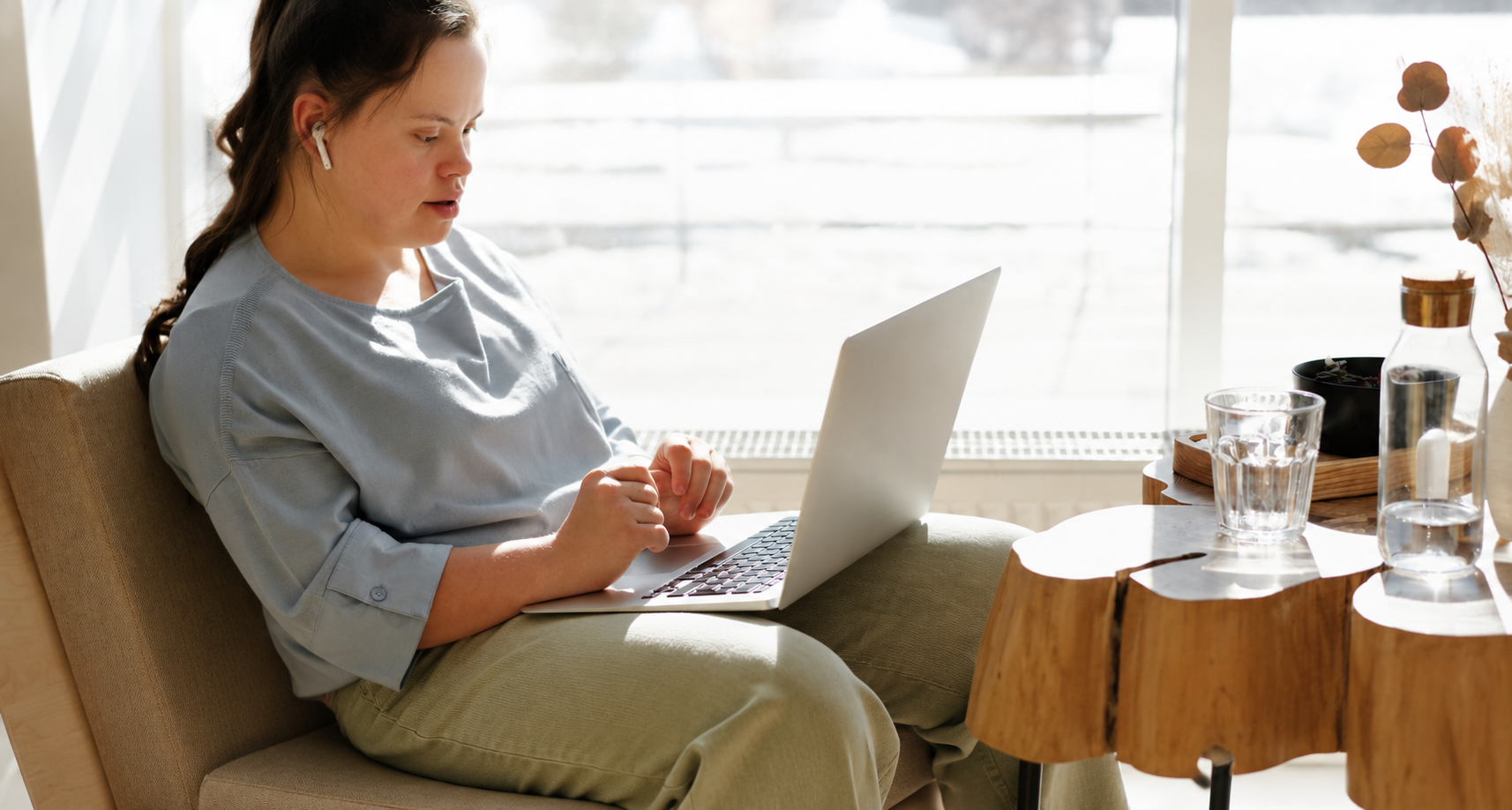 A young woman sitting in a sunlit armchair, working on a laptop at home.